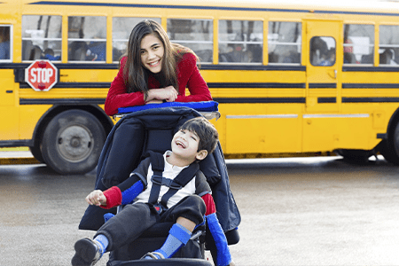 Big sister with disabled brother in wheelchair by school bus.